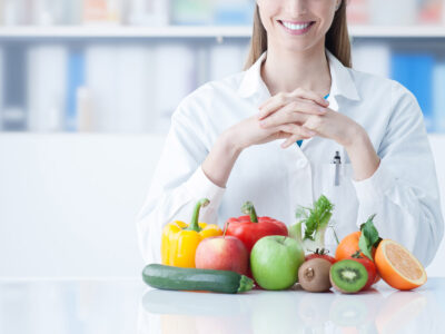 Smiling young dietician sitting at desk and showing colorful vegetables and fruit, healthy eating and diet concept