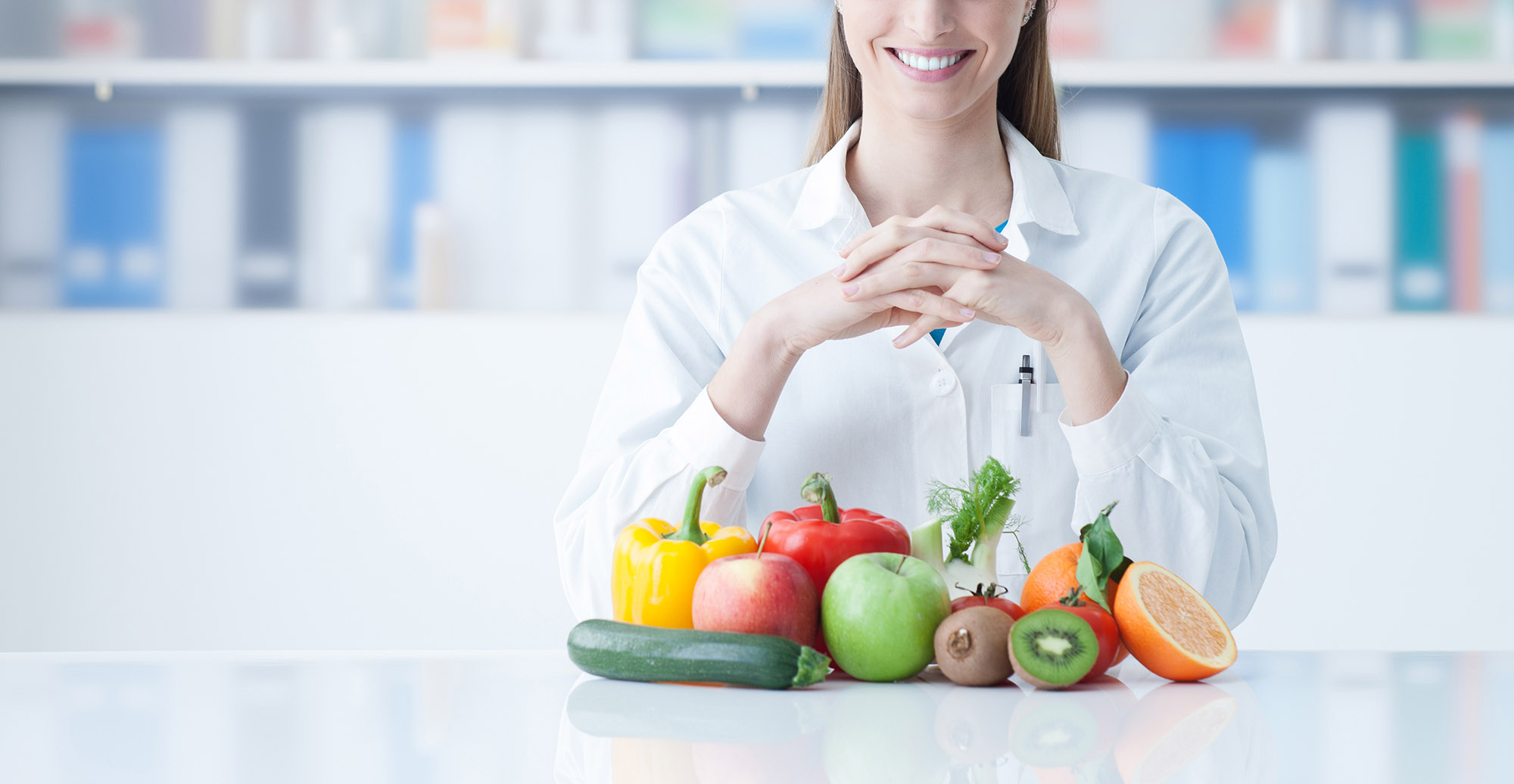 Smiling young dietician sitting at desk and showing colorful vegetables and fruit, healthy eating and diet concept